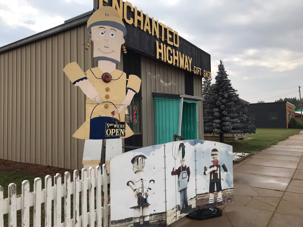 The exterior of the Enchanted Highway Gift Shop, a tan metal building with a black and gold sign, a life-sized cutout of a smiling farm woman figure, and a photo cutout board depicting three farm family figures, behind a white picket fence