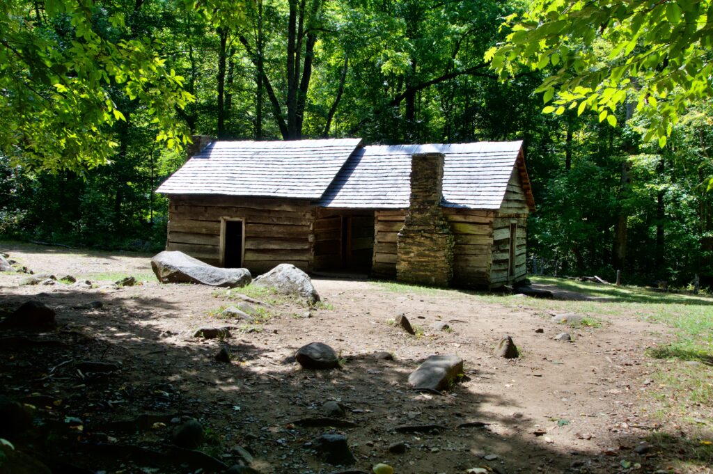 A historic "dog-trot" style log cabin with two connected structures under a shared roof and a stone chimney, set in a forest clearing along the Roaring Fork Motor Nature Trail
