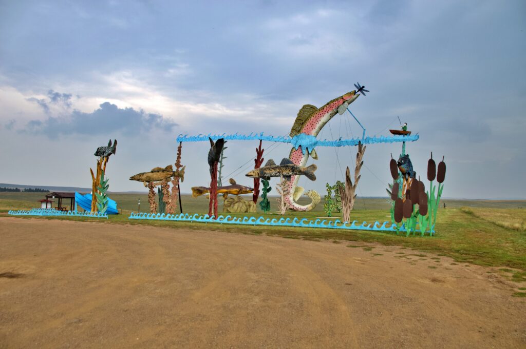 A large multi-part metal sculpture depicting several colorful fish leaping over a wavy blue metal border, including a giant pink and yellow trout jumping toward a dragonfly, with a small fisherman figure in a boat and cattail sculptures nearby, set on the open prairie