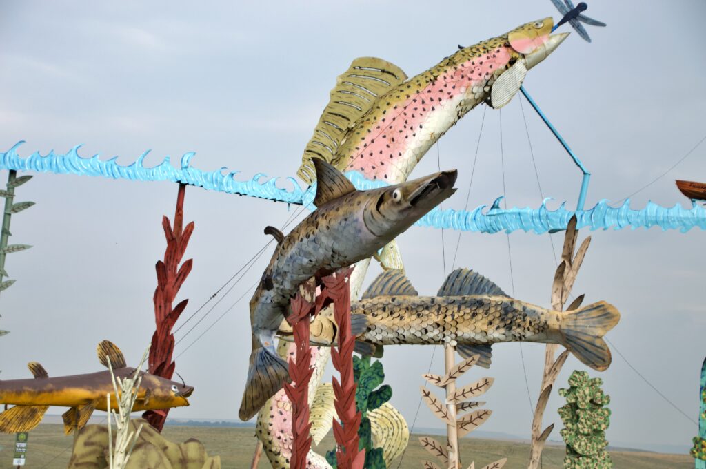 Close-up of several detailed metal fish sculptures leaping upward, including a pink and gold trout with individual scales reaching toward a metal dragonfly, layered in front of other fish, reeds, and cattail sculptures