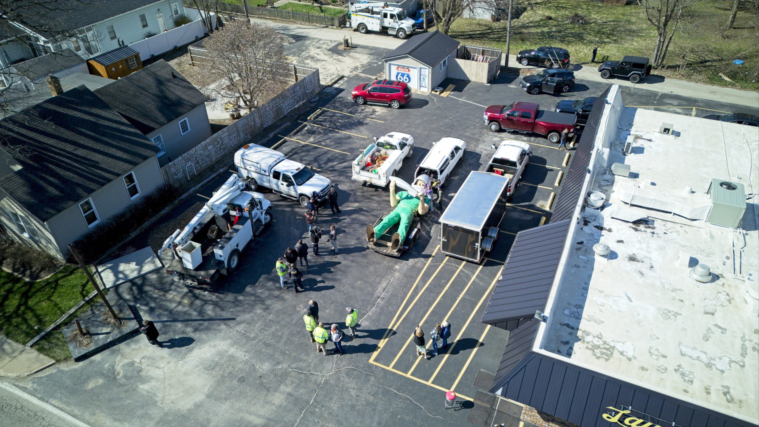 Aerial view of the Gemini Giant statue lying on its back on a flatbed trailer in a parking lot, surrounded by work crews and vehicles during its relocation