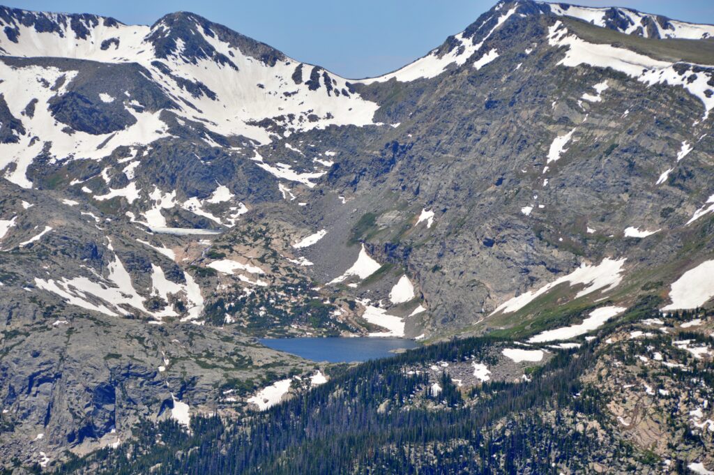 A distant alpine lake nestled at the base of steep, snow-streaked granite peaks, seen through a telephoto lens from a Trail Ridge Road overlook, with dense forest below and rocky, glaciated terrain above