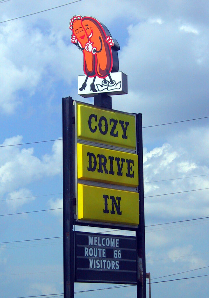 The Cozy Dog Drive-In sign along Route 66, topped with a cartoon logo of two hot dogs embracing, with a marquee reading "Welcome Route 66 Visitors"