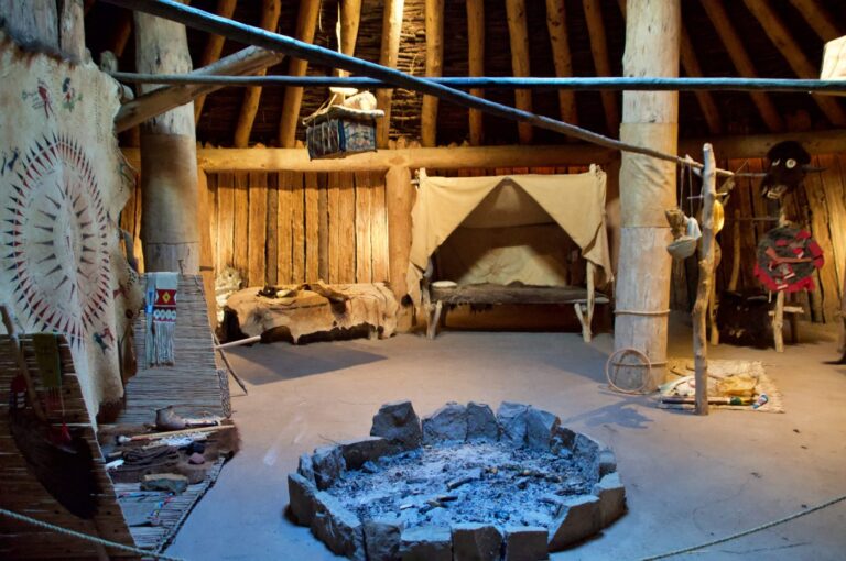 The interior of a reconstructed Hidatsa earth lodge at Knife River Indian Villages National Historic Site in Stanton, North Dakota. The circular living space features a central stone fire pit with ash, heavy log support posts, animal hide sleeping platforms, decorated painted hides, woven textiles, and traditional tools and implements arranged throughout. The domed log and earth roof structure is visible overhead. It was in villages like this one that Lewis and Clark spent the winter of 1804-05 and first met Sacagawea and her husband Toussaint Charbonneau