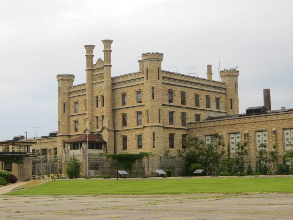 The historic Old Joliet Prison's castle-like limestone facade with turrets, arched windows, and razor-wire fencing under an overcast sky