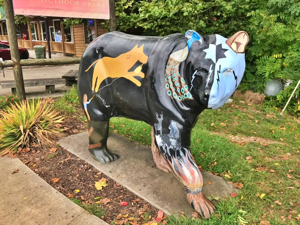 A black bear statue painted with scenes from Cherokee legends, including a horse, a deer, and a feathered headdress motif, standing outside a building near the Unto These Hills outdoor theater