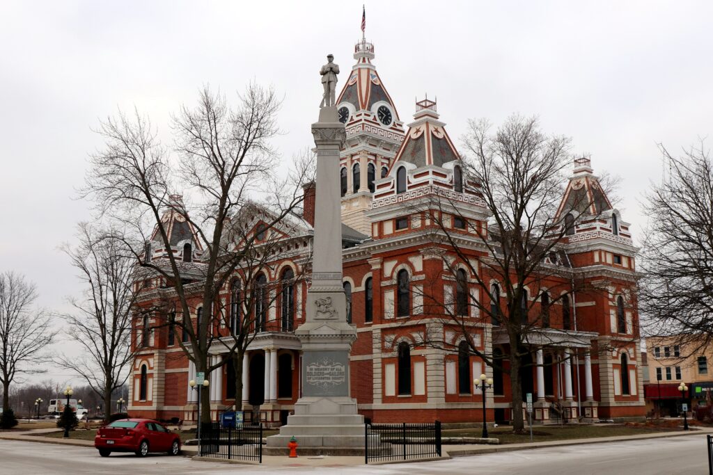 The ornate red brick and limestone Livingston County Courthouse with a clock tower and mansard roofs, framed by bare trees and a Civil War soldier memorial statue