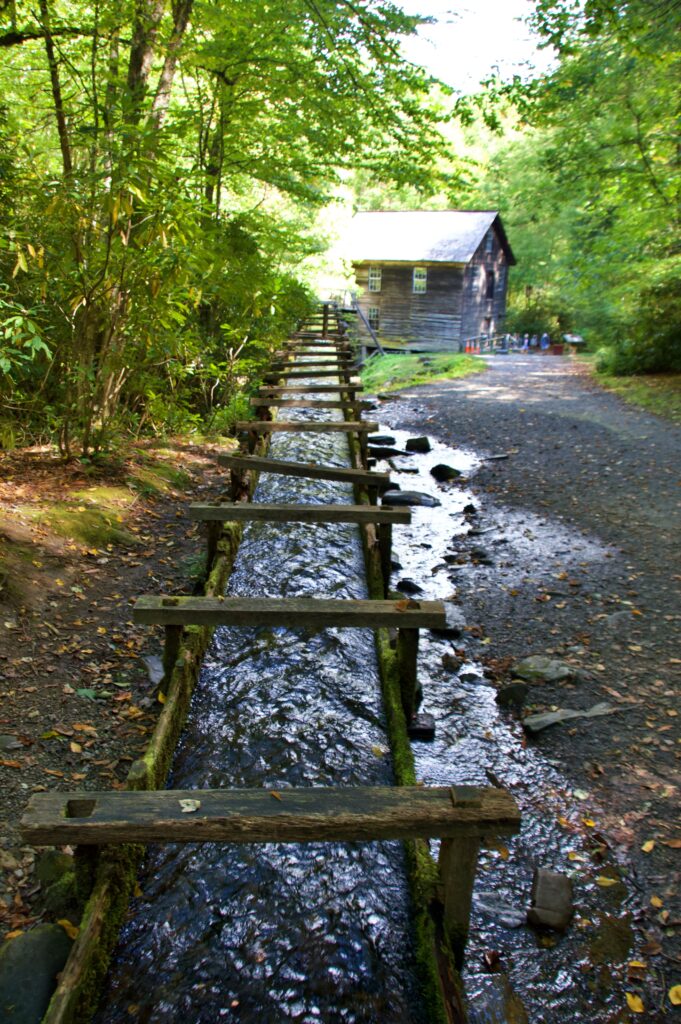 A wooden flume carrying water alongside a gravel path leading toward the weathered wooden Mingus Mill building, surrounded by forest