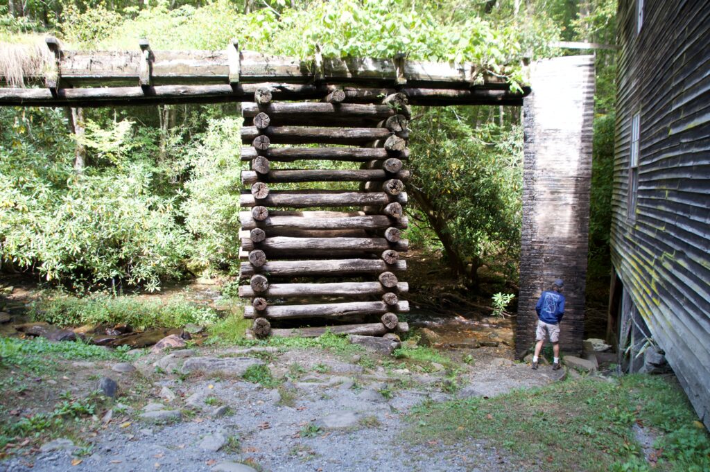 A man standing beside the tall wooden penstock of Mingus Mill, a stacked-log flume structure used to channel water down to the mill's turbine