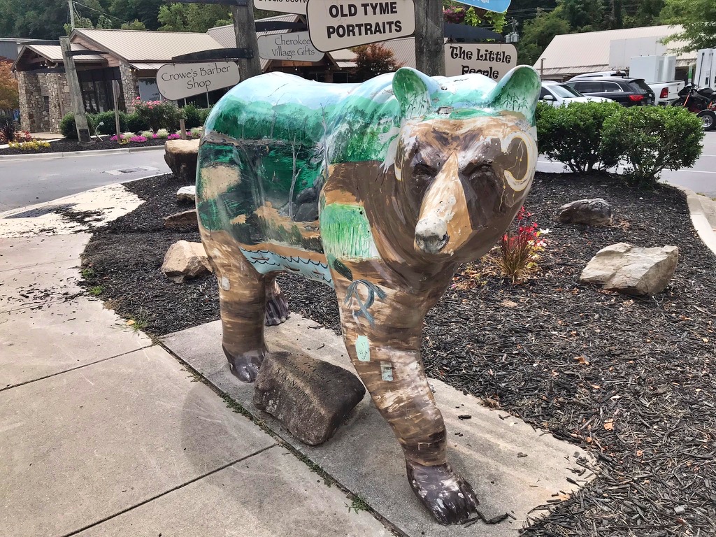 A bear statue painted with a forest and hibernation-den scene, part of Cherokee's painted bear public art trail, standing outside a row of downtown shops