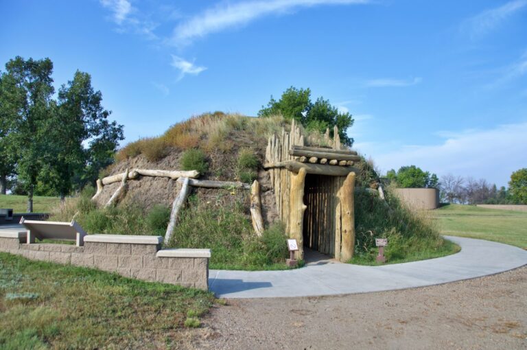 A reconstructed Hidatsa earth lodge at Knife River Indian Villages National Historic Site in Stanton, North Dakota. The dome-shaped structure is built from logs and covered with layers of earth and grass, with a wooden entry passage framing the doorway. A curved paved path leads visitors to the entrance. It was at this village complex that Lewis and Clark wintered in 1804-05 and hired interpreter Toussaint Charbonneau and his wife Sacagawea to join the expedition