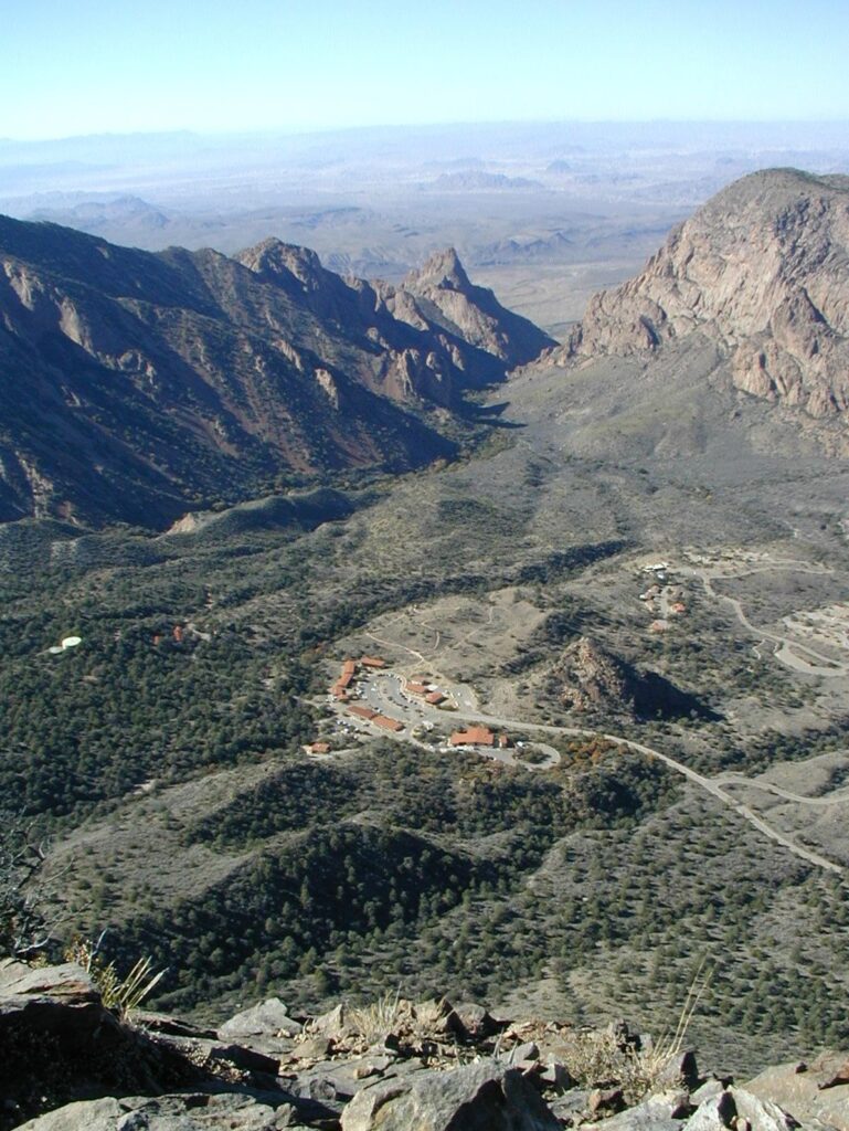 An aerial view looking down into the Chisos Basin, showing the lodge complex, Window Trail, and surrounding mountain peaks in Big Bend National Park