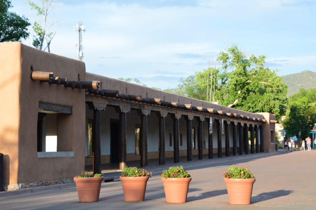 The long adobe facade of the Palace of the Governors in Santa Fe, with a traditional wooden portal, vigas, and terracotta planters lining the sidewalk