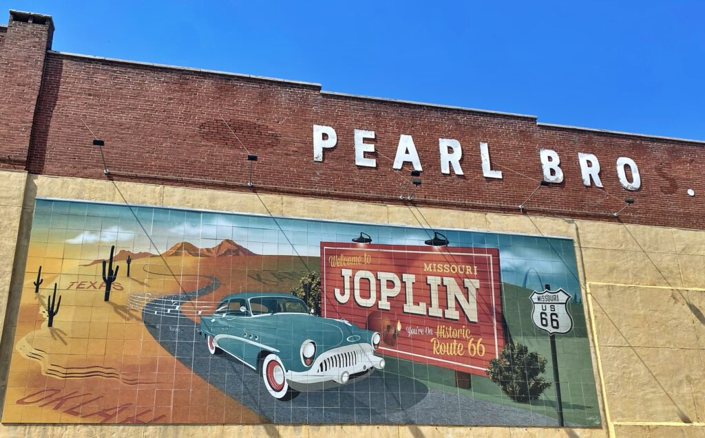 A tile mural on the side of a brick building painted with "Pearl Bros" lettering, depicting a teal vintage car driving toward a "Welcome to Joplin, Missouri" road sign, with a desert scene and cacti on the left