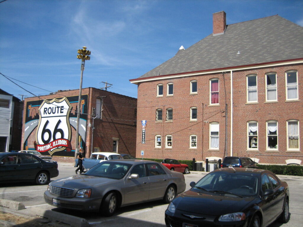 A Route 66 mural reading "Pontiac, Illinois" on a brick wall, with the historic Pontiac City Hall and Fire Station building visible nearby