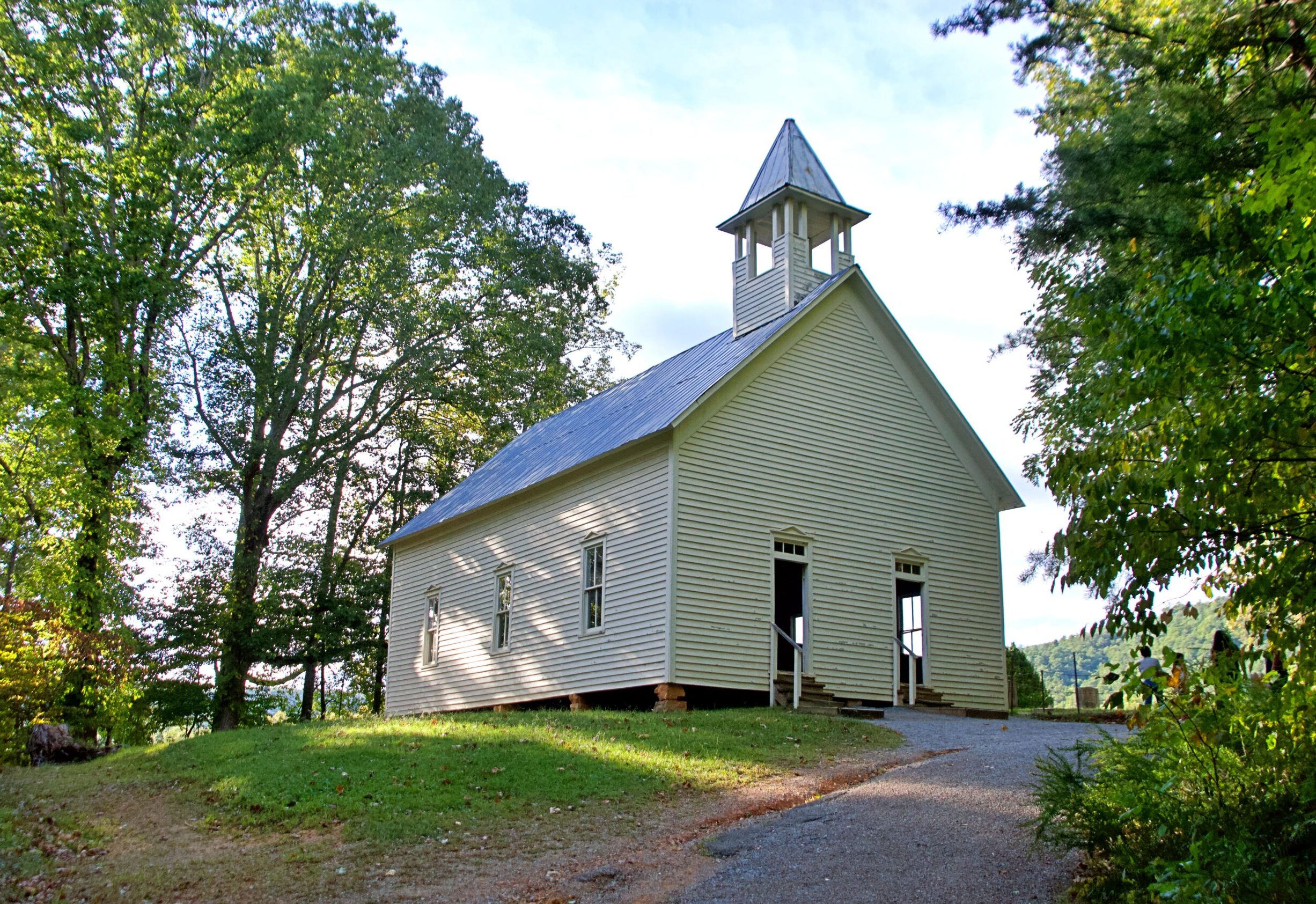 The Primitive Baptist Church at Cades Cove in Great Smoky Mountains National Park, Tennessee. The simple white clapboard church with a tin roof and small steeple sits on a gentle hillside surrounded by mature trees, with a gravel path leading to its open front door. Built in 1887, it is one of three historic churches preserved in Cades Cove