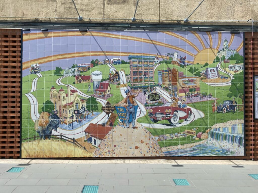 A large, colorful tile mural depicting a stylized map of Joplin, Missouri, with local landmarks including a bison, a train, historic buildings, a waterfall, and vintage cars, painted in a whimsical illustrated style