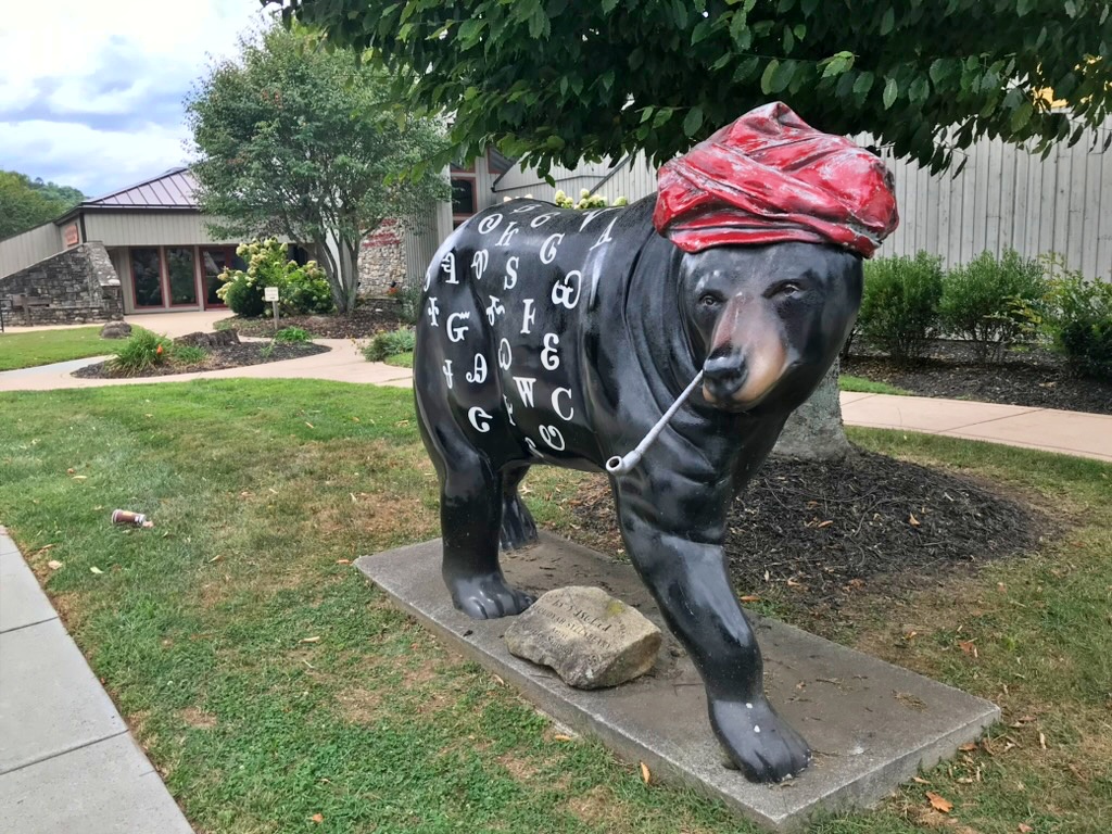 A black bear statue painted with Cherokee syllabary characters, wearing a red turban and holding a pipe, part of Cherokee's Great Bear Hunt trail