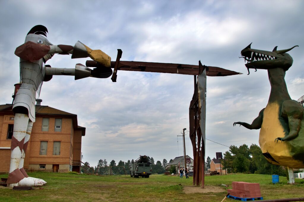 A giant metal knight sculpture named Sir Albert, wearing segmented mesh armor, thrusting a massive sword toward a fanged dragon sculpture across a grassy lawn, with a brick schoolhouse and water tower visible in the background for scale