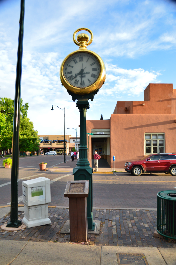 The Spitz Clock, a large gold pocket watch-shaped clock mounted on a green iron post, standing on a Santa Fe street corner near adobe buildings