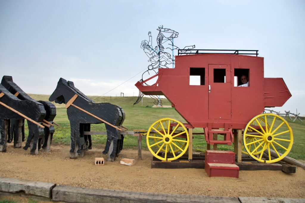A red wooden stagecoach with yellow spoked wheels and a team of black wooden horses, set up as a walk-in photo op, with the black wireframe Teddy Roosevelt sculpture visible on a grassy hill in the background