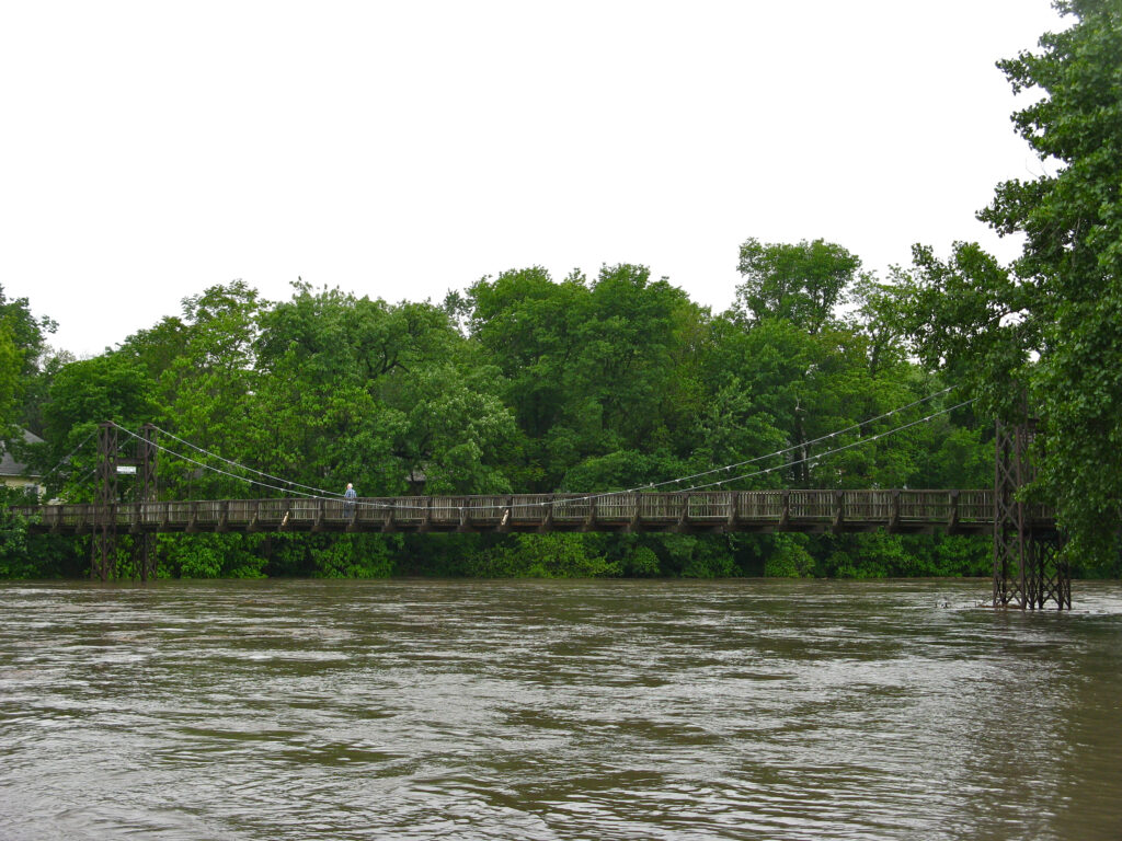 A wooden suspension footbridge crossing the wide, murky Vermilion River, with a person walking across and dense trees lining the far bank