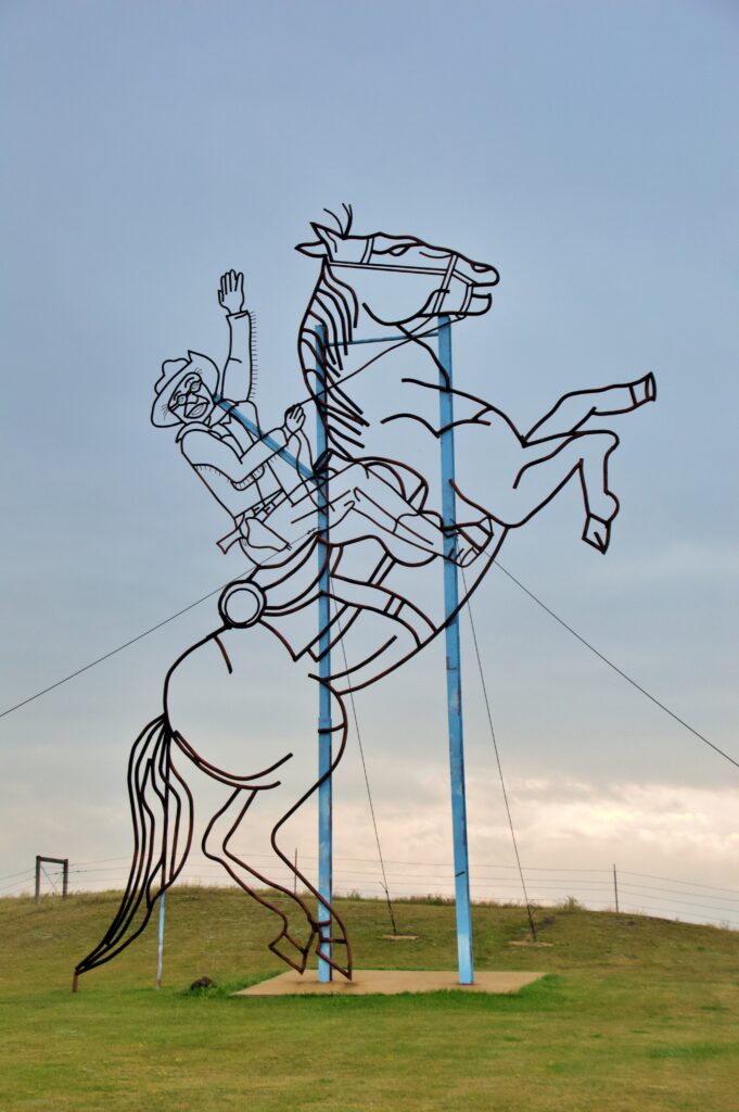 A tall black metal sculpture drawn in outline style depicting Theodore Roosevelt riding a rearing horse with one arm raised, set against a cloudy evening sky on an open grassy hill