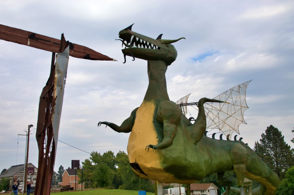 A large green and yellow metal dragon sculpture with an open fanged mouth, wing-like structures, and clawed feet, straining against a sword pointed at its throat, with houses and trees visible far below for scale