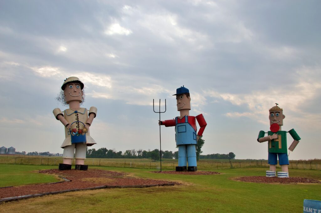 Three giant colorful metal figures representing a farm family — a mother in a dress holding a flower basket, a father in overalls holding a pitchfork, and a young son — standing on grass in a rural field