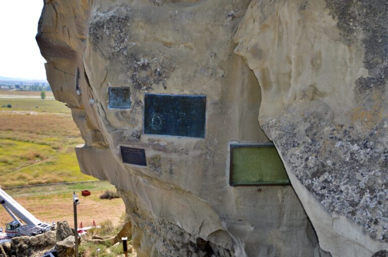 The sandstone face of Pompeys Pillar National Monument near Billings, Montana, showing the glass-protected case covering William Clark's carved signature from July 25, 1806, alongside several bronze memorial plaques mounted into the rock face. The vast Yellowstone River valley stretches across the background far below, with farm fields and the town of Billings visible in the distance. The green-tinted glass case on the right protects the only verified original physical evidence of the Lewis and Clark Expedition remaining in place on the entire 4,900-mile trail