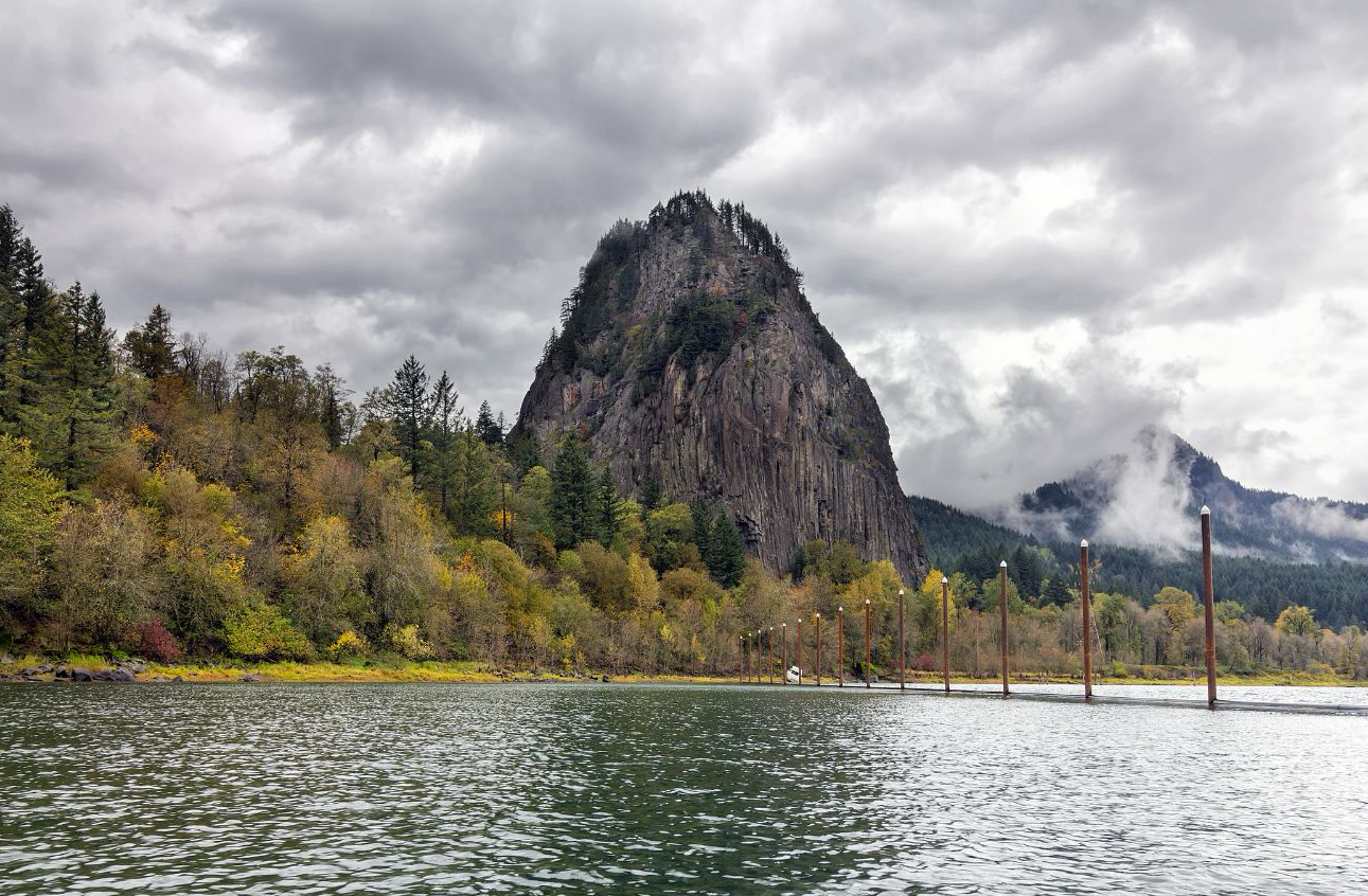Beacon Rock rising above the Columbia River in Washington State, with autumn foliage, misty mountains, and marina pilings in the foreground. A landmark on the Lewis and Clark National Historic Trail visited by the Corps of Discovery in October 1805