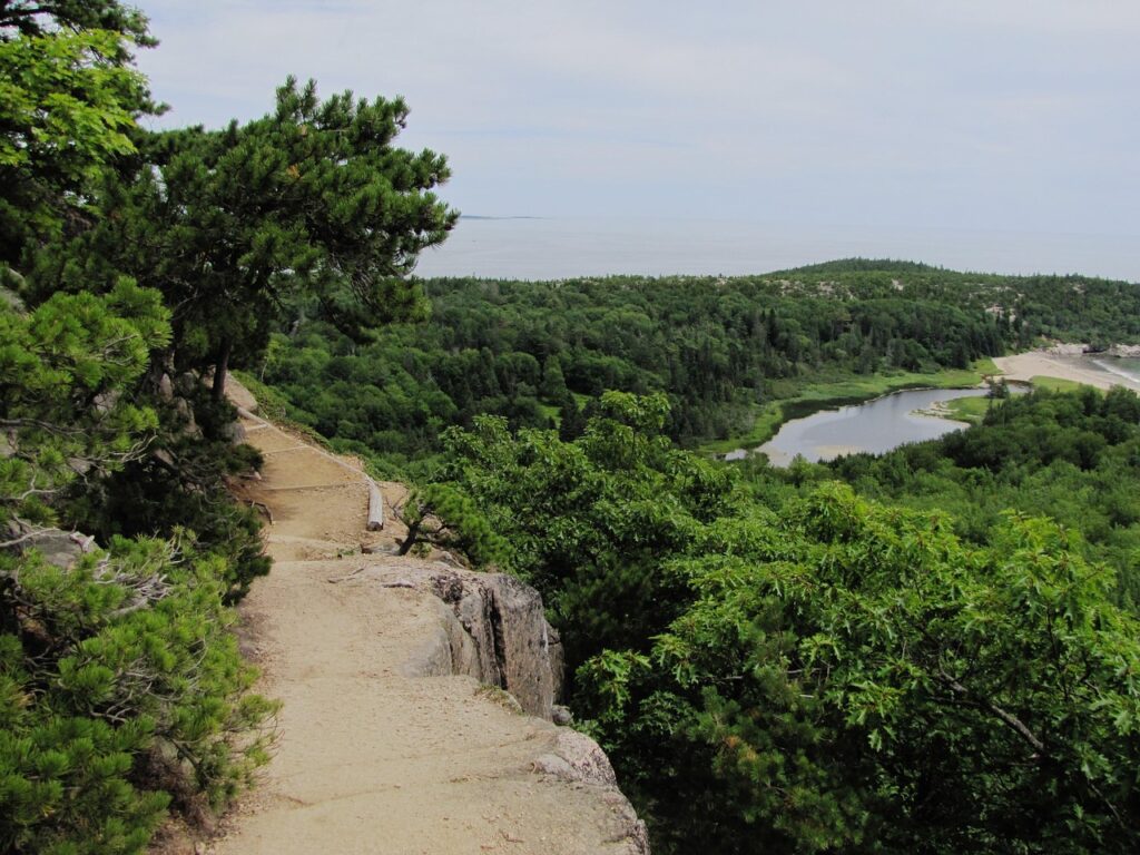 A hiking trail along an exposed cliffside with a coastal pond and forest visible below in Acadia National Park