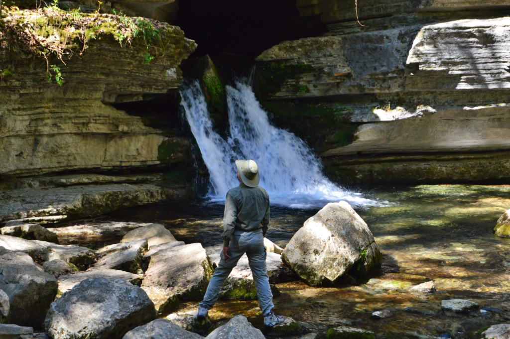 Hiker in a wide-brimmed hat standing before Blanchard Spring emerging from a cave opening at Blanchard Springs Caverns, Arkansas