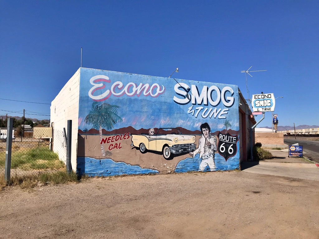 A colorful mural on the side of an auto shop in Needles, California, showing a vintage yellow convertible, a palm tree, and an Elvis-style figure, with "Econo Smog & Tune" painted above