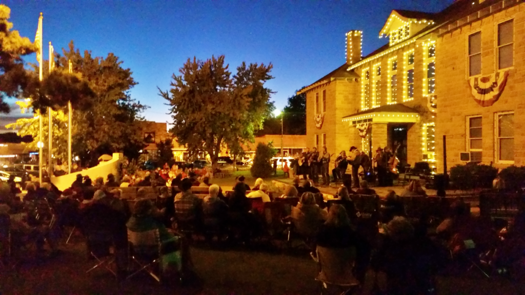Live folk music performance on the Mountain View town square at dusk, with the illuminated Stone County Courthouse in the background"