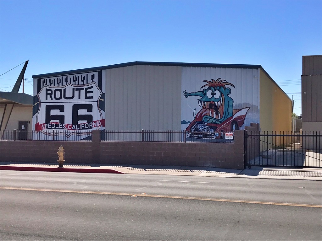 cartoon mural of a wide-eyed hot-rod driver painted on a building wall in Needles, California, beneath a large Route 66 shield reading "Needles California"