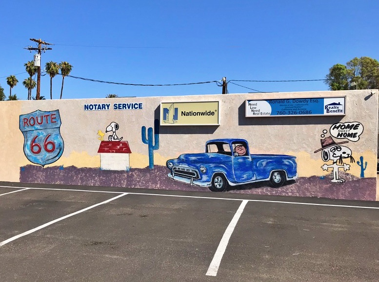 A mural in Needles, California, showing Snoopy on his doghouse beside a Route 66 shield, with a blue pickup truck and a 'Home Sweet Home' speech bubble painted alongside
